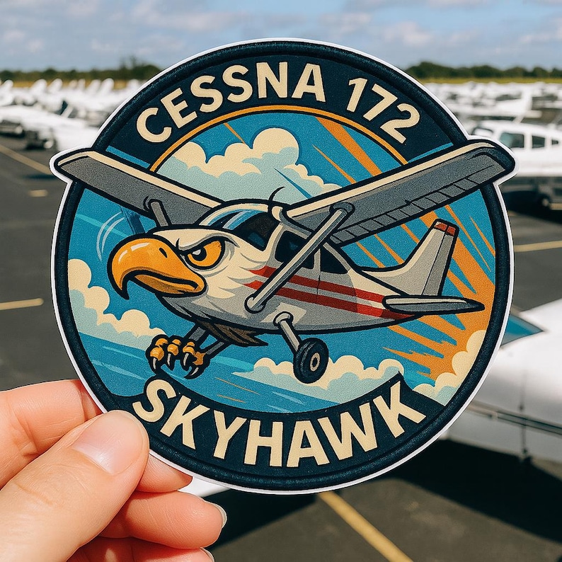 May include: A circular sticker featuring a cartoon Cessna 172 Skyhawk airplane with an eagle head. The design includes the text "CESSNA 172 SKYHAWK" in white and yellow against a blue and orange background. The sticker is held by a hand.