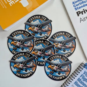 May include: A collection of Cessna 172 Skyhawk stickers featuring an eagle graphic, surrounded by a blue and orange design. The stickers are placed near aviation study guides and manuals, including a "Federal Aviation Administration" book.