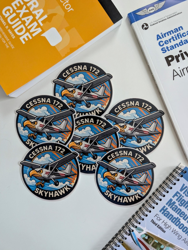 May include: A collection of Cessna 172 Skyhawk stickers featuring an eagle graphic, surrounded by a blue and orange design. The stickers are placed near aviation study guides and manuals, including a "Federal Aviation Administration" book.