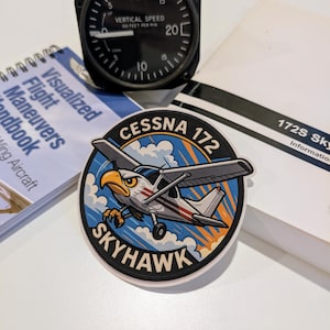 May include: A black vertical speed indicator gauge with white markings and a needle pointing to 10. A sticker with a cartoon Cessna 172 Skyhawk airplane and eagle graphic. A flight maneuvers handbook and a 172S Skyhawk information booklet are also present.