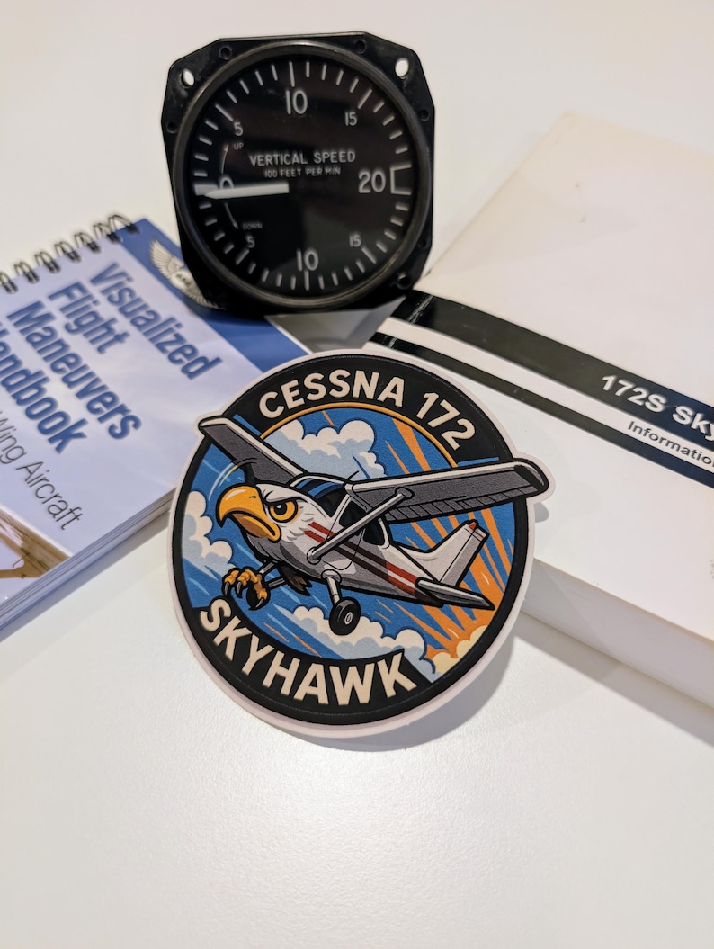 May include: A black vertical speed indicator gauge with white markings and a needle pointing to 10. A sticker with a cartoon Cessna 172 Skyhawk airplane and eagle graphic. A flight maneuvers handbook and a 172S Skyhawk information booklet are also present.