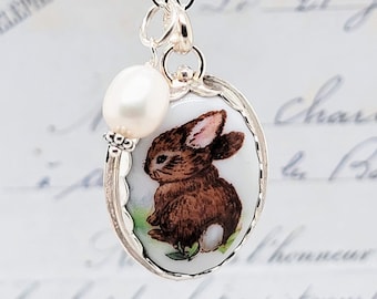 May include: A silver necklace featuring an oval pendant with a painted brown rabbit on a white background. The pendant is framed in scalloped silver and is accented by a single white oval pearl charm hanging from the same silver chain link.