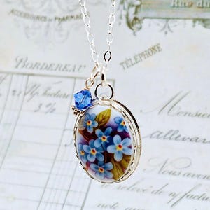 May include: A silver chain necklace featuring an oval pendant with a floral design of blue forget-me-not flowers on a white background, accented by a small, faceted, deep blue crystal bead attached to the top of the pendant frame.