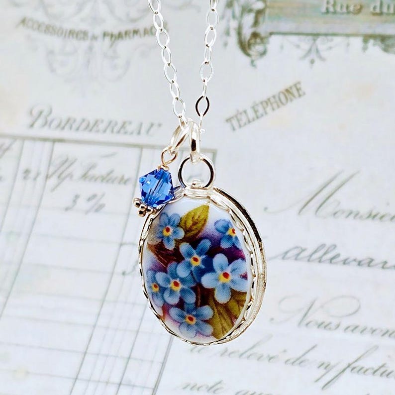 May include: A silver chain necklace featuring an oval pendant with a floral design of blue forget-me-not flowers on a white background, accented by a small, faceted, deep blue crystal bead attached to the top of the pendant frame.