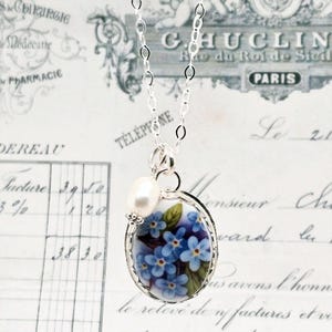 May include: A silver chain necklace featuring an oval pendant with a blue forget-me-not floral pattern and a single white freshwater pearl accent. The pendant is framed in silver and hangs from a delicate silver link chain.