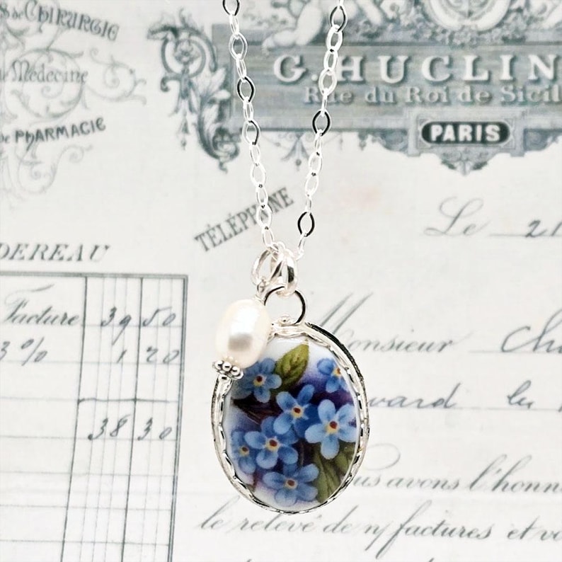 May include: A silver chain necklace featuring an oval pendant with a blue forget-me-not floral pattern and a single white freshwater pearl accent. The pendant is framed in silver and hangs from a delicate silver link chain.