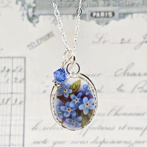 May include: A silver chain necklace featuring an oval pendant with a painted floral design of blue forget-me-not flowers and green leaves. A small, faceted blue crystal bead is attached to the top of the silver-toned metal pendant frame.