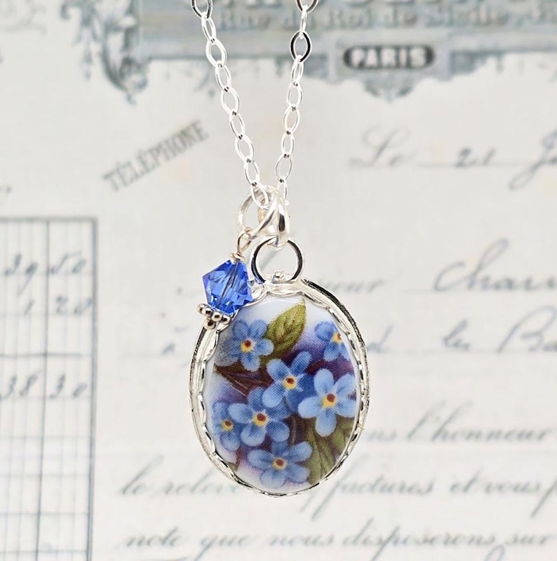 May include: A silver chain necklace featuring an oval pendant with a painted floral design of blue forget-me-not flowers and green leaves. A small, faceted blue crystal bead is attached to the top of the silver-toned metal pendant frame.