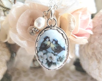 May include: A silver-toned oval pendant necklace featuring a painted illustration of two small birds perched on white flowers. The pendant is suspended from a silver chain and accented with a single white pearl bead attached to the top bail.