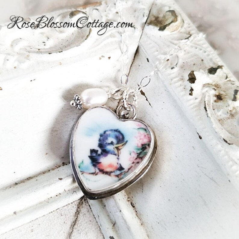 K&ouml;nnte beinhalten: Eine herzf&ouml;rmige Silberanh&auml;nger-Halskette mit einer farbenfrohen Illustration eines kleinen blauen Vogels, der auf einem Ast mit rosa Bl&uuml;ten sitzt. Der Anh&auml;nger ist mit einer einzelnen wei&szlig;en Perle verziert und h&auml;ngt an einer feinen Silberkette.
