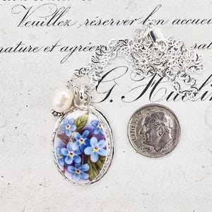 May include: A silver necklace featuring an oval pendant with a blue forget-me-not floral design and a small white pearl accent. A US dime is placed next to the pendant for scale, showing the relative size of the jewelry piece against the coin.