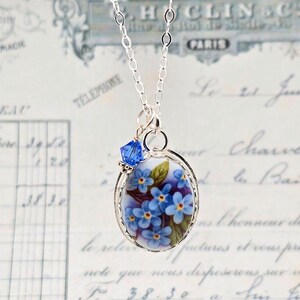 May include: A silver chain necklace featuring an oval pendant with a floral design of blue forget-me-not flowers and green leaves on a white background, accented by a single small faceted blue crystal bead attached to the top jump ring.