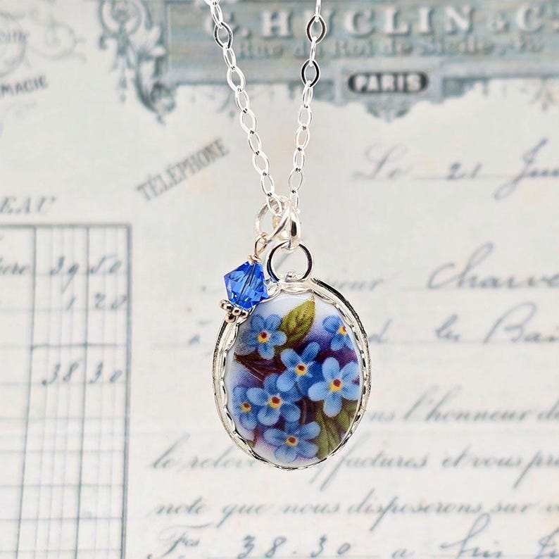 May include: A silver chain necklace featuring an oval pendant with a floral design of blue forget-me-not flowers and green leaves on a white background, accented by a single small faceted blue crystal bead attached to the top jump ring.