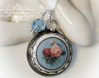 May include: A silver pendant necklace featuring a round ceramic insert with a floral design of pink and red roses on a light blue background, framed by a scalloped silver border, accompanied by a single faceted light blue crystal bead charm.