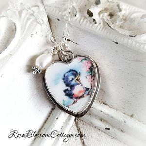 K&ouml;nnte beinhalten: Eine herzf&ouml;rmige Anh&auml;ngerkette mit einer wei&szlig;en Keramikbasis, die eine farbenfrohe Illustration eines kleinen blauen Vogels und rosa Blumen zeigt. Der Anh&auml;nger ist in silbernes Metall gefasst und h&auml;ngt an einer Kette neben einer wei&szlig;en Perle.