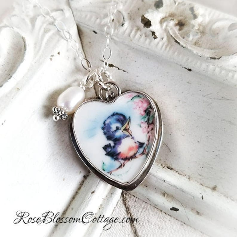 K&ouml;nnte beinhalten: Eine herzf&ouml;rmige Anh&auml;ngerkette mit einer wei&szlig;en Keramikbasis, die eine farbenfrohe Illustration eines kleinen blauen Vogels und rosa Blumen zeigt. Der Anh&auml;nger ist in silbernes Metall gefasst und h&auml;ngt an einer Kette neben einer wei&szlig;en Perle.