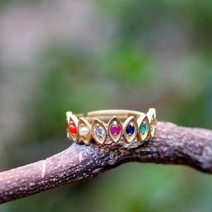 May include: A gold ring with a series of colourful gemstones. The ring features red, white, clear, pink, blue, and green stones set in a repeating pattern. The ring is displayed on a brown branch against a blurred green background.