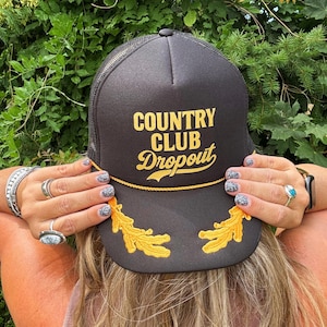 May include: Black trucker hat with the text "COUNTRY CLUB DROPOUT" in gold lettering. The hat features a gold rope detail and two gold leaf designs. The hat is worn by a person with blonde hair.