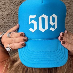 May include: A bright turquoise trucker hat with the number "509" in bold white lettering. The hat has a curved brim and a mesh back. The hat is being held up by a person.