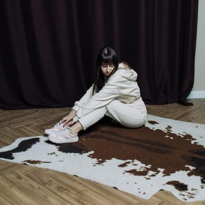 May include: A cream-colored hooded sweatshirt and matching sweatpants set. The person is sitting on a cowhide rug with brown and white patches. The person is wearing pink and white sneakers. The background features a dark brown curtain and a light-colored wall.