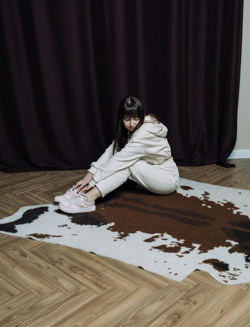May include: A cream-colored hooded sweatshirt and matching sweatpants set. The person is sitting on a cowhide rug with brown and white patches. The person is wearing pink and white sneakers. The background features a dark brown curtain and a light-colored wall.