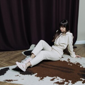 May include: A beige tracksuit consisting of a hooded zip-up jacket and matching pants. The outfit is paired with pink and white sneakers. The person is sitting on a cowhide rug with brown, black, and white patches.