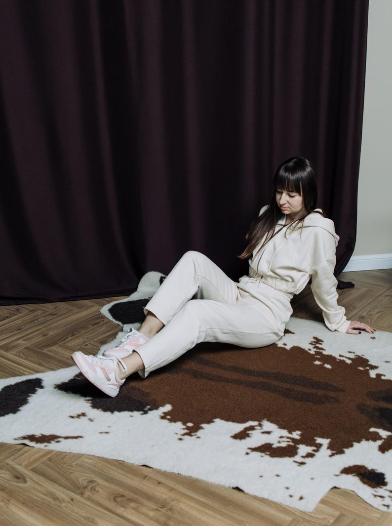 May include: A beige tracksuit consisting of a hooded zip-up jacket and matching pants. The outfit is paired with pink and white sneakers. The person is sitting on a cowhide rug with brown, black, and white patches.