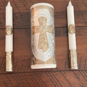 May include: A unity candle set with two white taper candles and a central pillar candle. The pillar candle and the base of the taper candles have gold and silver cross designs. The candles are on a dark wood surface.