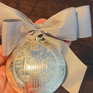 May include: A silver-colored medallion with an image of a saint and Latin text, attached to a light gray ribbon bow. The medallion has intricate details and is suspended from the bow.