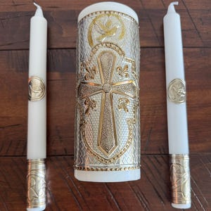 May include: A white unity candle set with two taper candles. The center candle is silver and gold with a cross and dove design. The taper candles have gold accents and seals. The set is on a wooden surface.