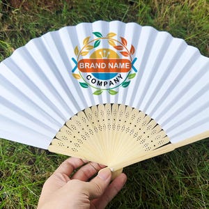 Pode incluir: Um leque de papel branco dobrável com um logótipo decorativo. O leque está aberto, mostrando o seu design plissado. A pega é feita de madeira de cor clara. O logótipo inclui as palavras "BRAND NAME COMPANY" num design circular com folhas.