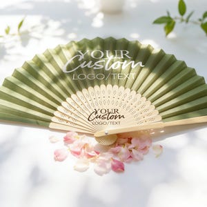 Peut inclure: Un éventail pliable avec un tissu vert olive et une structure en bois clair. Le tissu présente le texte blanc "YOUR Custom LOGO/TEXT" et les baleines en bois portent le texte noir "YOUR Custom LOGO/TEXT" avec des motifs décoratifs découpés.