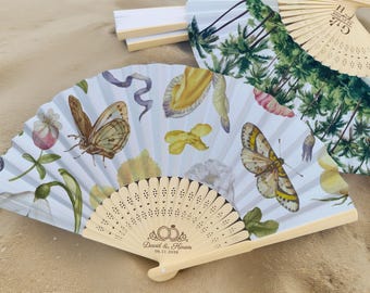 May include: A collection of decorative hand fans with floral and butterfly designs. One fan features a light wood handle with the names "David & Karen" and the date "06.11.2026" engraved. The fans are set on a sandy surface.