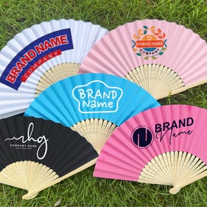 Pode incluir: Uma coleção de leques de papel abertos em várias cores, incluindo branco, rosa, azul e preto, com cabos de madeira. Cada leque apresenta um nome de marca e um design de logotipo diferente. Os leques estão dispostos sobre relva verde.