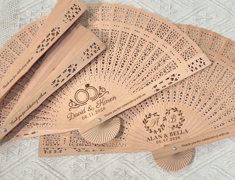 May include: Several light brown wooden fans with intricate cut-out designs are displayed. Some fans feature personalized text with names and dates, along with the phrase "Thank you for celebrating with us."