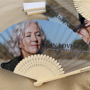 May include: A wooden folding fan with a printed image of a woman with the text "IN LOVING memory Megan Smith 1967-2025" and "Forever in my heart". The fan is light beige with a light blue background.