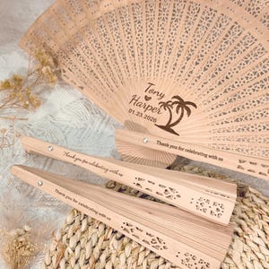 May include: Wooden hand fans with intricate cut-out designs. One fan is fully open, displaying the names "Tony Harper" and the date "01.23.2026" with a palm tree illustration. The fans are engraved with the message "Thank you for celebrating with us."