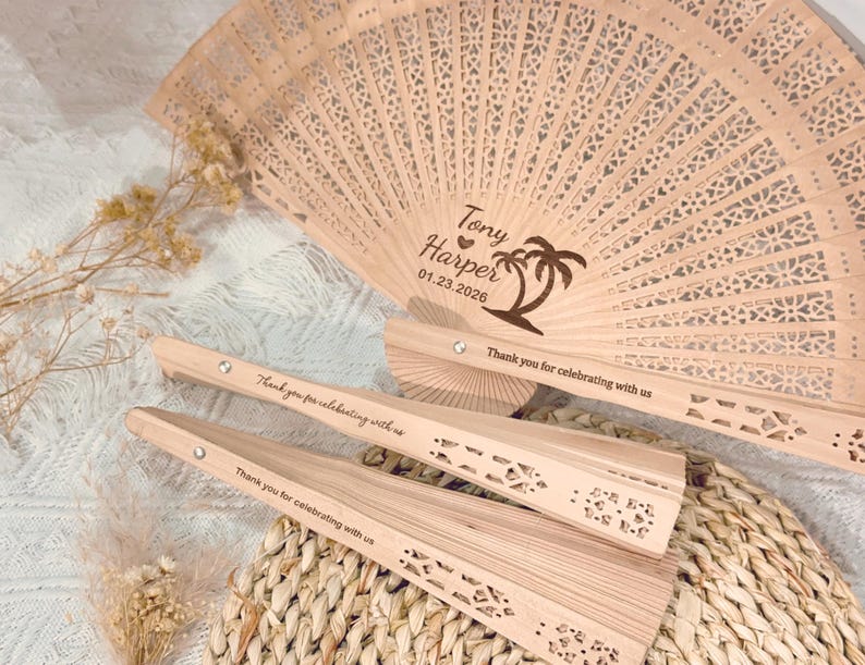 May include: Wooden hand fans with intricate cut-out designs. One fan is fully open, displaying the names "Tony Harper" and the date "01.23.2026" with a palm tree illustration. The fans are engraved with the message "Thank you for celebrating with us."