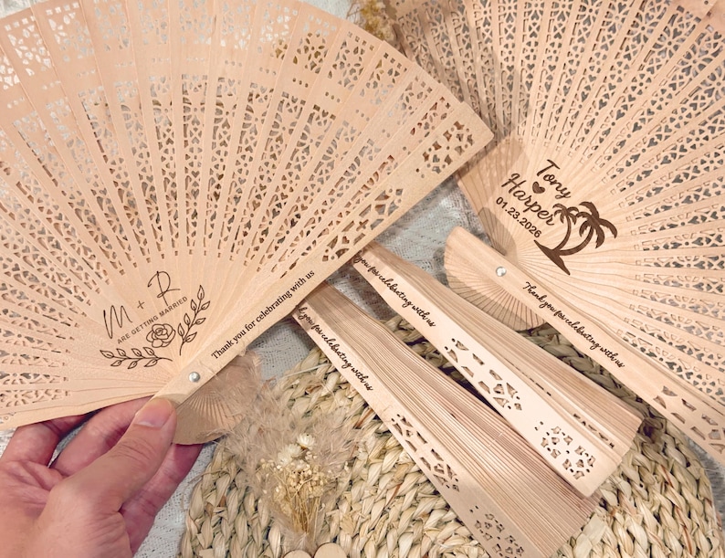 May include: Wooden fans with intricate cut-out designs. One fan displays "M + R ARE GETTING MARRIED" with floral accents. Another fan has "Tony Harper 01.23.2026" and a palm tree. All fans have "Thank you for celebrating with us" text.