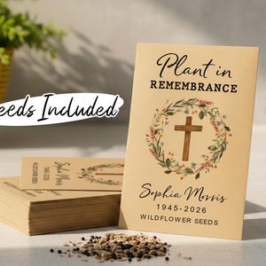 Può includere: Confezioni di semi di fiori selvatici commemorativi con la scritta "Plant in Remembrance" e "Sophia Morris 1945-2026". Le confezioni presentano una croce circondata da una ghirlanda floreale. Sono visibili anche le parole "Seeds Included".
