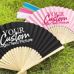 Może przedstawiać: Dwa otwarte wachlarze, jeden czarny i jeden różowy, z napisem "YOUR Custom Logo/Text/Picture". Wachlarze wykonane są z papieru i bambusa. Inne wachlarze w kolorze niebieskim, różowym i białym są w tle.