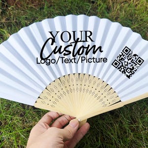 May include: White folding fan with the text "YOUR Custom Logo/Text/Picture" and a QR code. The fan has a light beige wooden handle and is open, displaying its pleated design. The fan is held against a background of green grass.