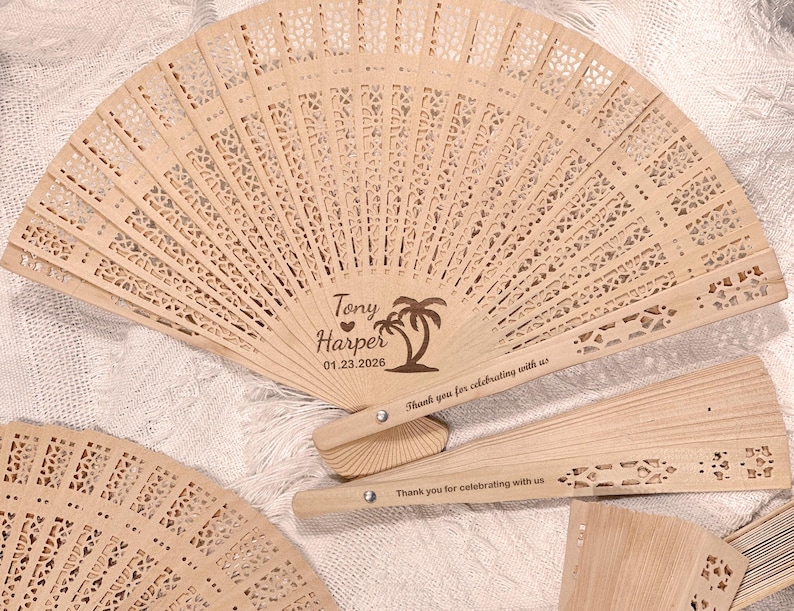 May include: Several open, light-colored wooden fans with intricate cut-out designs are displayed. One fan is open, displaying the names "Tony Harper" and the date "01.23.2026" with a palm tree graphic. The text "Thank you for celebrating with us" is also visible.