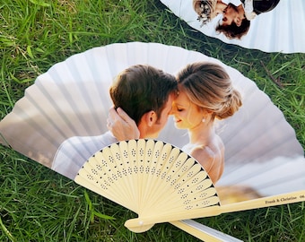 May include: A pair of wedding fans with a photograph of a couple embracing. The fans are made of light-colored wood and paper, with a decorative pattern of small cut-out shapes. The fans are lying on green grass.