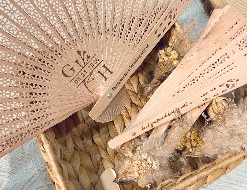 May include: Two light brown wooden fans with intricate cut-out designs. One fan has the initials "G H" and the date "03.21.2026" engraved. The text "Thank you for celebrating with us" is printed on the fan. The fans are in a woven basket.