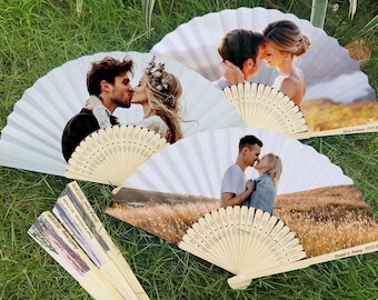 May include: White paper fans with images of couples kissing, set against a backdrop of green grass. The fans have wooden handles and are personalized with names and dates. A unique wedding favor.
