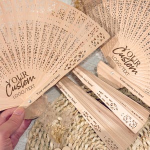 Peut inclure: Plusieurs éventails en bois clair avec des motifs ajourés complexes. Les éventails portent l'inscription "YOUR Custom LOGO/TEXT". Le texte "Your custom text here!" est également visible sur les manches des éventails.