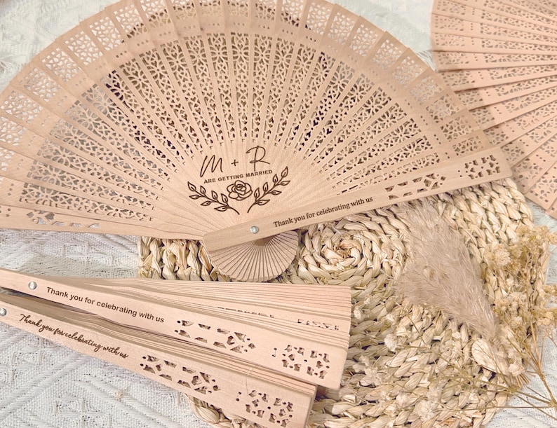 May include: Several open wooden fans with intricate cut-out designs are displayed. The fans are light brown and feature the text "M+R ARE GETTING MARRIED" and "Thank you for celebrating with us." The fans are arranged on a woven mat.