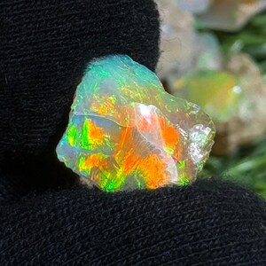 May include: A vibrant, raw opal gemstone with a spectrum of colors including green, orange, and yellow. The opal is set against a dark background, highlighting its iridescent qualities. The rough-cut stone displays a natural, unpolished texture.