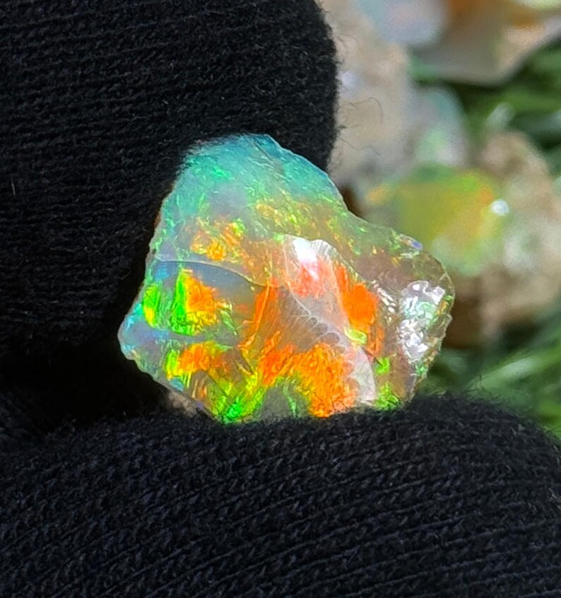 May include: A vibrant, raw opal gemstone with a spectrum of colors including green, orange, and yellow. The opal is set against a dark background, highlighting its iridescent qualities. The rough-cut stone displays a natural, unpolished texture.